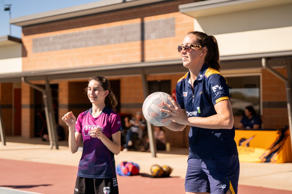 Lightning player Cara Koenen holds a netball up as if she is about to pass it. There is a young player next to her who is smiling. 